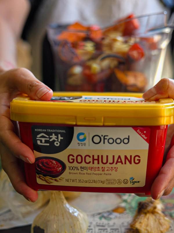 A person holding a container of OFood Korean Traditional Gochujang, a brown rice red pepper paste, with visible garlic bulbs below. In the background is a glass bowl with chopped vegetables.